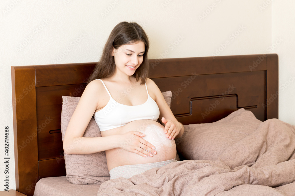 a pregnant girl sits at home on the bed and smears an anti-stretch mark cream on her stomach. Pregnancy, motherhood, preparation and expectation concept a pregnant girl sits at home on the bed and smears an anti-stretch mark cream on her stomach. Pregnancy, motherhood, preparation and expectation concept