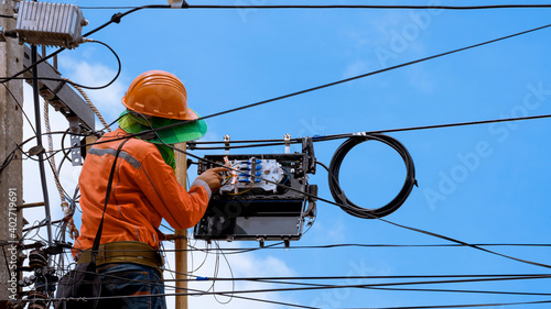 Rear view of technician on wooden ladder checking fiber optic cables in internet splitter box on electric pole against blue sky background photo