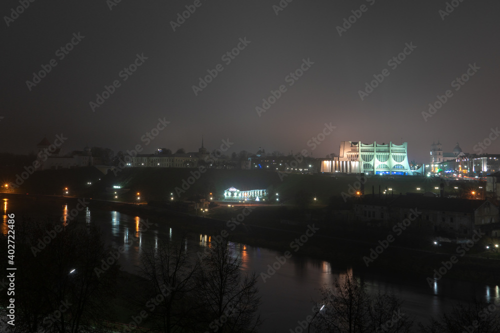 Night panorama of the big city at the foot of the river. Lots of colorful burning lights and street lighting. Top view of houses in fog at night.