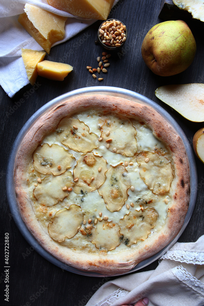Pizza with dried pears and pine nuts. Traditional Italian pizza. Metal plate for serving pizza. A piece of parmesan and a pear on a dark background. White cotton napkin. The view from the top. Pizza with dried pears and pine nuts. Traditional Italian pizza. Metal plate for serving pizza. A piece of parmesan and a pear on a dark background. White cotton napkin. The view from the top.