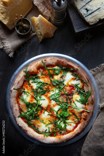 Pizza with several types of cheese, arugula and sauce. Traditional Italian pizza. Metal plate for serving pizza. Piece of cheese on a dark background. Vertical photo. View from above.