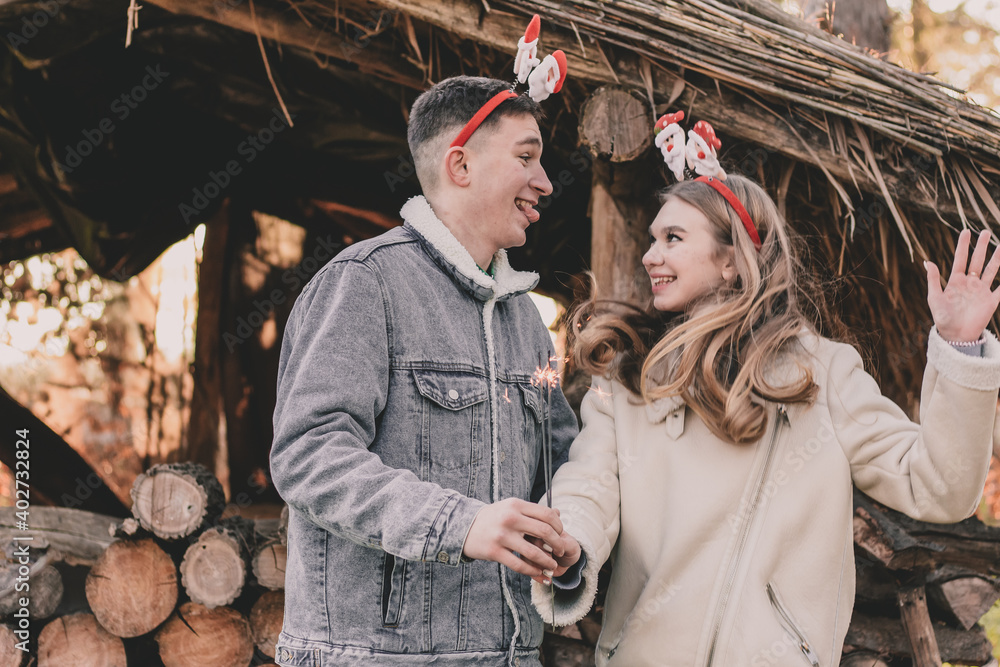 A couple about their appearance are dressed in New Year's hoops on their heads, holding a sparkler in their hands and smiling at each other against the background of a gazebo made of firewood