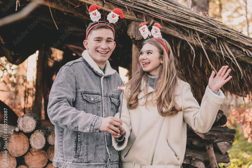 A couple about their appearance, dressed in New Year's hoops on their heads, holds a sparkler in their hands and smiles against the background of a gazebo made of firewood