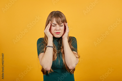Headache grimacing pain holds the back of neck indicating location. Fatigue during workaholism labor. Young attractive woman, dressed green shirt blonde hair, yellow background
