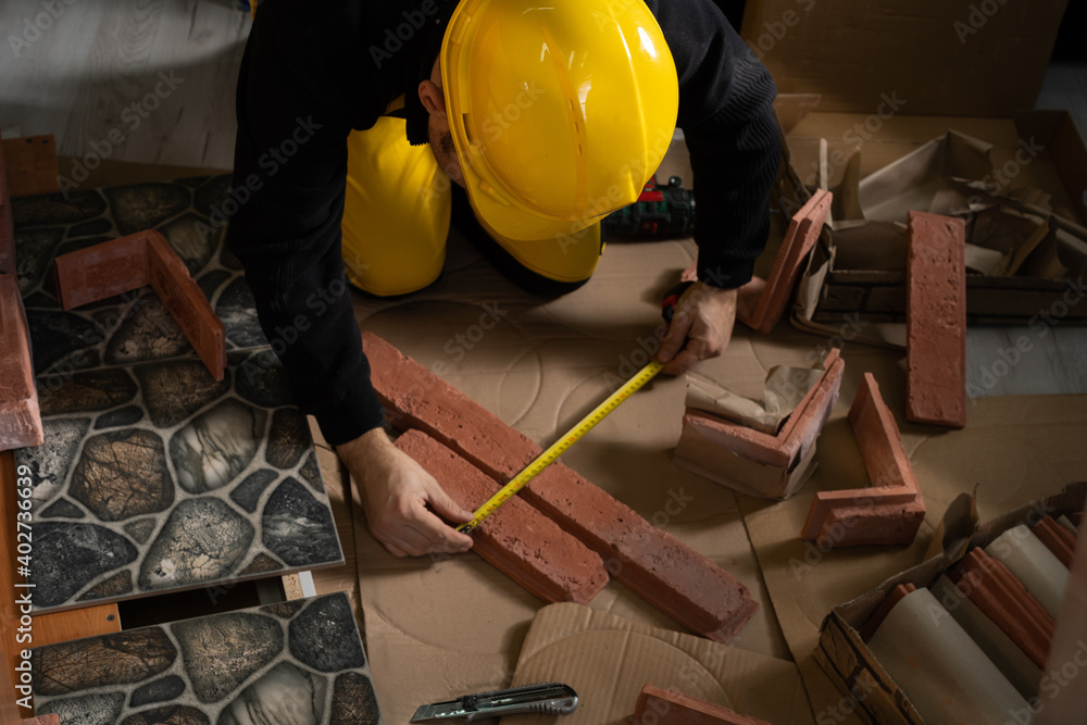 A construction worker in working clothes, kneeling, measures the retractable decorative tiles in the shape of old red brick with a tape measure. A construction worker in working clothes, kneeling, measures the retractable decorative tiles in the shape of old red brick with a tape measure.