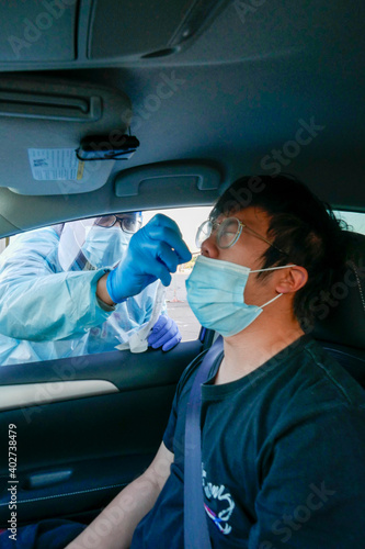 Medical personnel administering rapid COVID-19 test with  a long nasal swab to a young Asian man inside a car at a drive-thru testing site