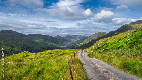 Long country road leading to majestic Black Valley with lake and mountain range covered in sunlight and shadows from clouds, County Kerry, Ireland