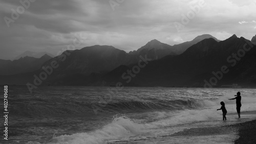 Silhouette of two kids enjoying the wavy sea on a rainy day in Antalya, Turkey. Black and white photograph of children, sea, mountains and clouds.