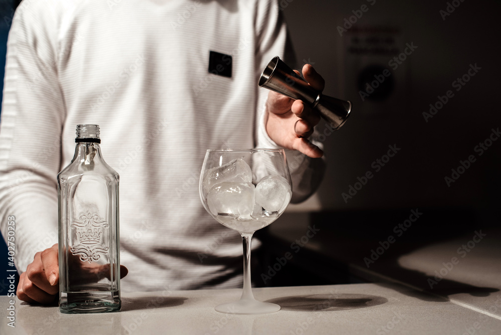 Un camarero joven vertiendo un alcohol blanco transparente en una copa con hielos sobre la barra de un bar usando un medidor Un camarero joven vertiendo un alcohol blanco transparente en una copa con hielos sobre la barra de un bar usando un medidor