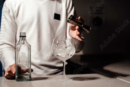 Un camarero joven vertiendo un alcohol blanco transparente en una copa con hielos sobre la barra de un bar usando un medidor