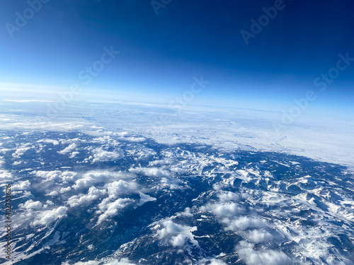 An aerial view from an airplane window of mountains, snow, clouds and blue skies.