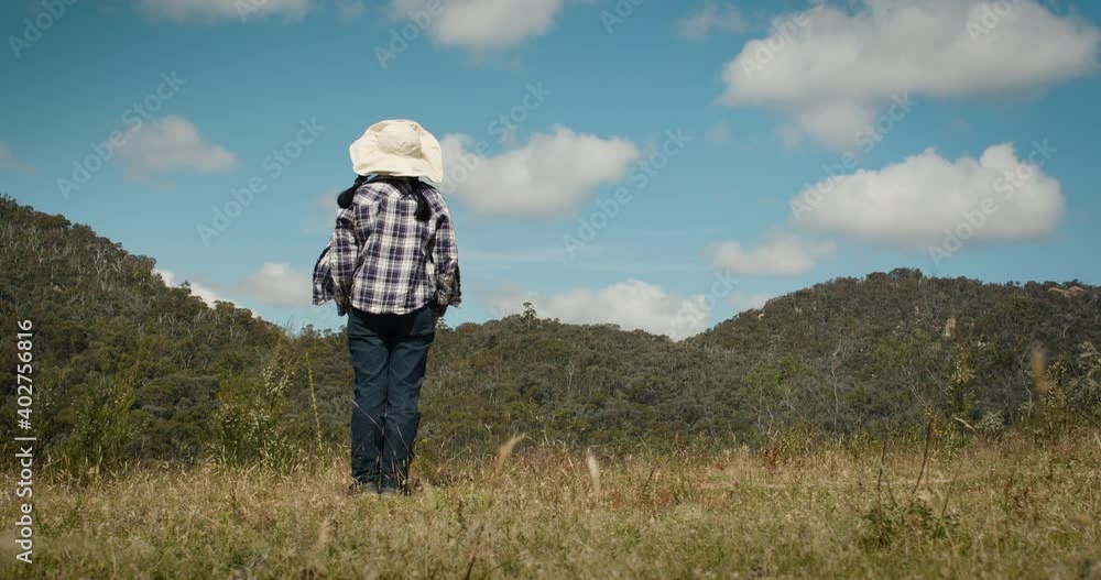 Young girl wearing a checked long-sleeve shirt, jeans and white hat standing on dry brown grass looking at forest covered hills with blue sky above, exploring wilderness located in Australia. Young girl wearing a checked long-sleeve shirt, jeans and white hat standing on dry brown grass looking at forest covered hills with blue sky above, exploring wilderness located in Australia.