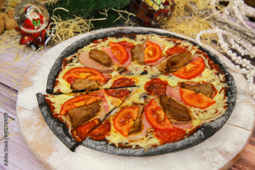Italian black dough pizza served on wooden plate and table with Christmas new year decoration on background. Fast food meat dish with pork, beef, tomato slices and mozzarella cheese 