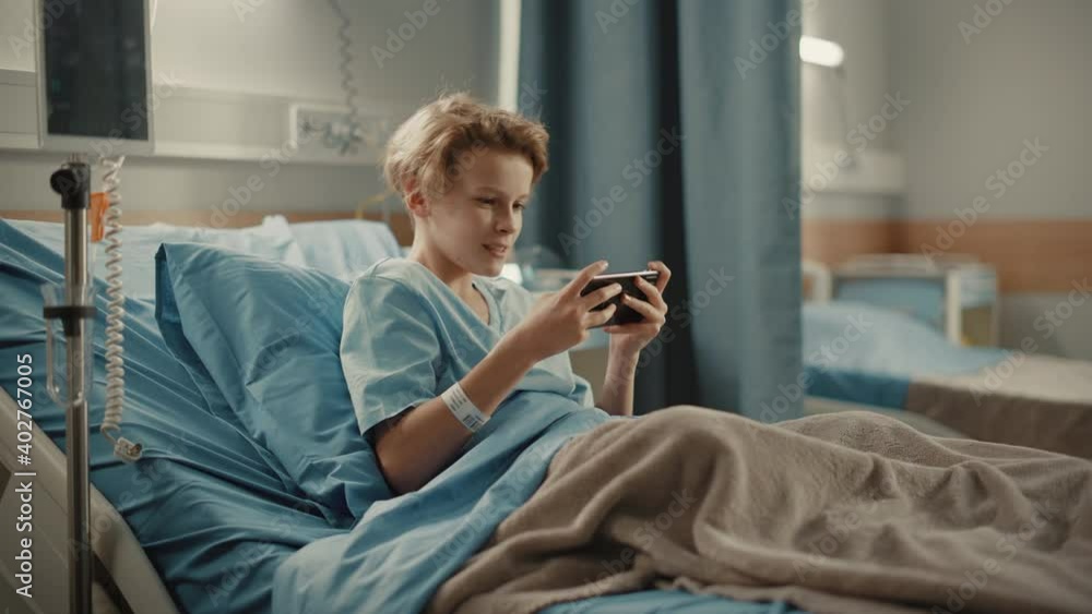 Hospital Ward: Handsome Young Boy Resting in Bed, Uses Smartphone, Plays Online Video Games on Internet with Mobile Phone. Happy Patient Recovering after Sickness Hospital Ward: Handsome Young Boy Resting in Bed, Uses Smartphone, Plays Online Video Games on Internet with Mobile Phone. Happy Patient Recovering after Sickness