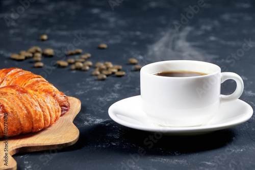 A cup of hot coffee and croissants on a black background. Breakfast with coffee and fresh pastry. Coffee beans on a table. Fresh cake and pastry.