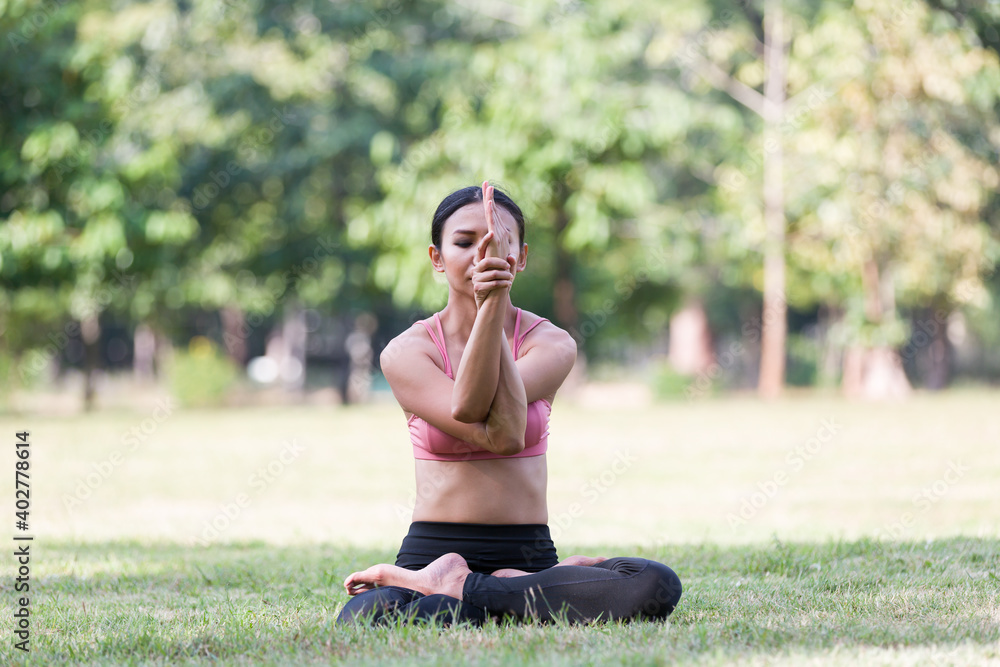 Fototapeta premium Asian woman doing yoga with stretching arms while sitting on green grass outdoor in the park. Woman wearing sportswear and working out outdoor. Yoga and exercise outdoor concept
