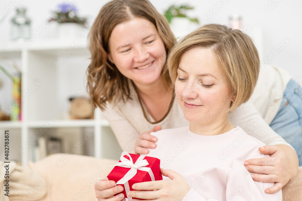 Two young women are hugging at home on the couch with a bright gift in their hands. Gay lesbian couple at home. Lesbian couple concept Two young women are hugging at home on the couch with a bright gift in their hands. Gay lesbian couple at home. Lesbian couple concept