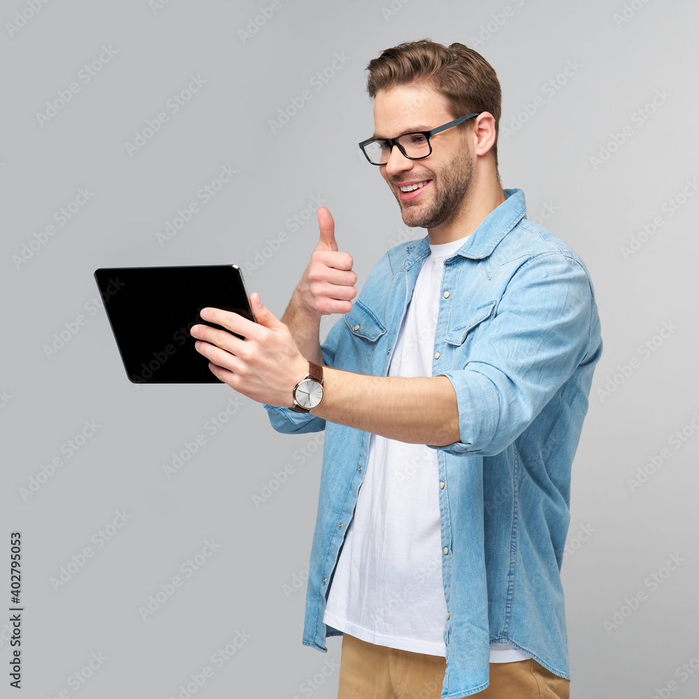 Happy young man wearing jeans shirt standing and using tablet over studio grey background Happy young man wearing jeans shirt standing and using tablet over studio grey background