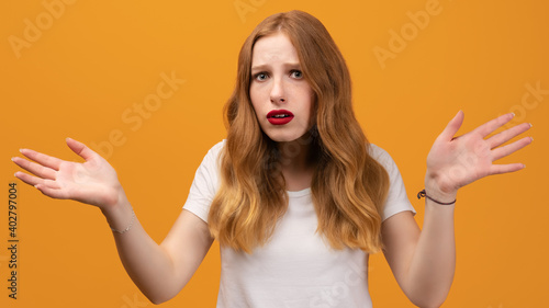 Puzzled girl with long wavy blonde hair, wearing white t-shirt spreads hands with doubt