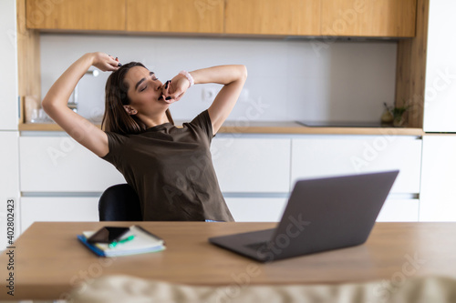 Young tired woman sitting at kitchen chatting by laptop and yawning.
