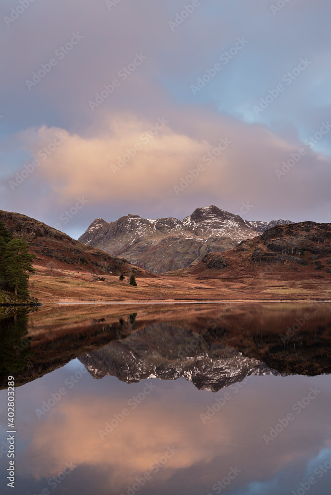 Beautiful vibrant Winter sunrise over Blea Tarn in Lake District with snow capped Langdale Pikes in distance