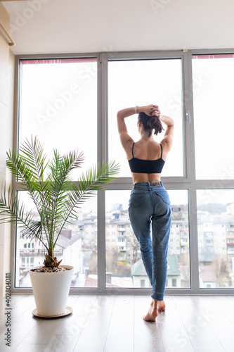 Rear view beautiful young woman standing with hands outstretched in bedroom, looking out large panoramic window in modern apartment, starting new day, enjoying morning