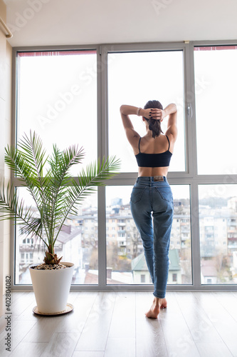 Good morning. Back view of happy young woman stretching and looking out the window at home
