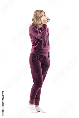 Side view of young woman in comfy home clothing drinking coffee or tea. Full body length isolated on white background.