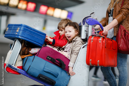 Two little kids, boy and girl, siblings and mother at the airport. Children, family traveling, going on vacation by plane and waiting on trolley with suitcases pushing by woman at terminal for flight.