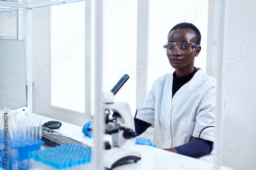 Woman of african ethnicity working in healthcare facility sitting at her workplace with protective glasses. Black healthcare scientist in biochemistry laboratory wearing sterile equipment.