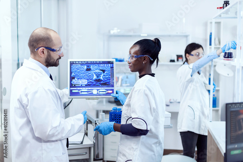 African assitant discussing wtih scientist about virus experiment in chemistry laboratory. Multiethnic team of medical researchers working together in sterile lab wearing protection glasses and gloves