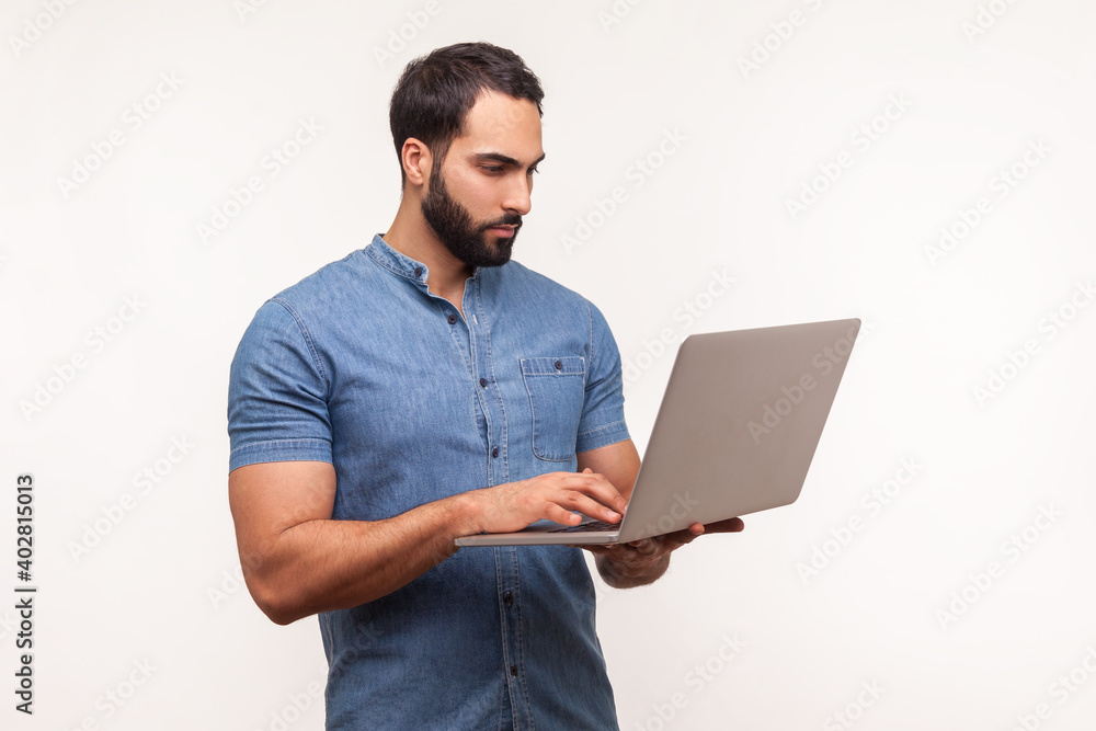Self confident serious man with beard in blue shirt holding wireless computer in hand, working on laptop, making edits, deadline. Indoor studio shot isolated on white background Self confident serious man with beard in blue shirt holding wireless computer in hand, working on laptop, making edits, deadline. Indoor studio shot isolated on white background