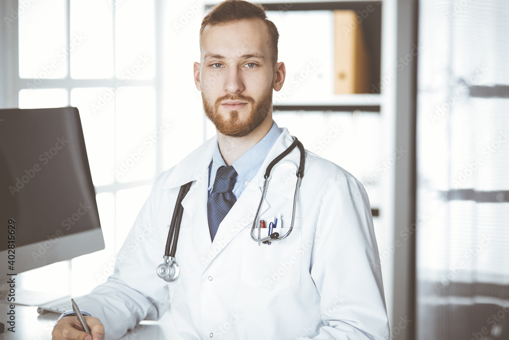 Friendly red-bearded doctor sitting and writing at clipboard in clinic. Perfect medical service in hospital. Medicine concept Friendly red-bearded doctor sitting and writing at clipboard in clinic. Perfect medical service in hospital. Medicine concept