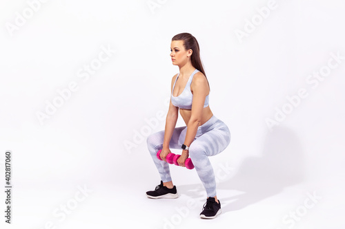 Side view full length assertive sportive woman in white top and tights doing squat exercises with dumbbell, pumping body muscles, developing strength. Indoor studio shot isolated on gray background
