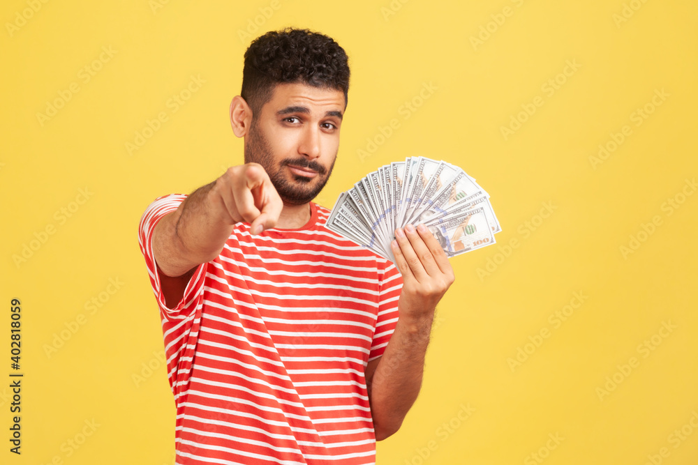 Self confident rich man with beard in striped t-shirt holding fan of dollars and pointing finger at camera with serious expression, easy profit. Indoor studio shot isolated on yellow background Self confident rich man with beard in striped t-shirt holding fan of dollars and pointing finger at camera with serious expression, easy profit. Indoor studio shot isolated on yellow background