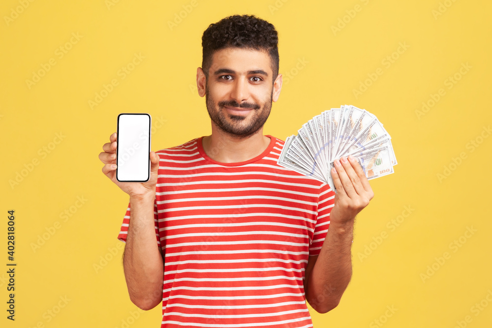 Smiling positive bearded man in striped t-shirt showing hundred dollar bills and smartphone with white display, bragging with online profit, banking app. Indoor studio shot isolated yellow background Smiling positive bearded man in striped t-shirt showing hundred dollar bills and smartphone with white display, bragging with online profit, banking app. Indoor studio shot isolated yellow background
