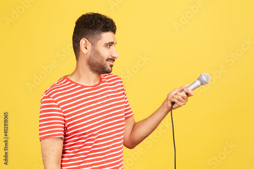 Profile portrait bearded man journalist in striped red t-shirt holding microphone, asking questions, discussing problems, interviewing. Indoor studio shot isolated on yellow background