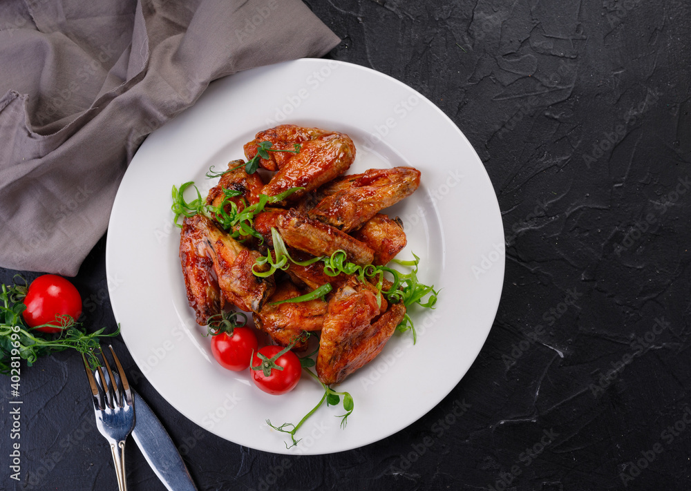barbecue chicken wings in a white plate on a dark background. Buffalo wings decorated with greens and cherry tomatoes barbecue chicken wings in a white plate on a dark background. Buffalo wings decorated with greens and cherry tomatoes