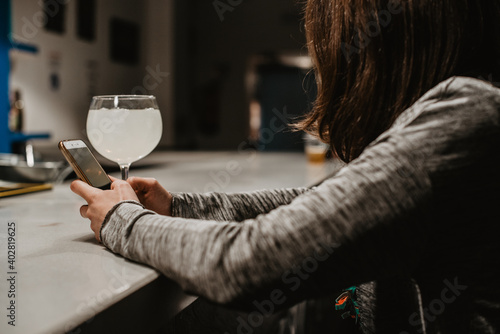 Una mujer joven sentada sola en la barra de un bar sosteniendo y mirando su teléfono móvil. En el mostrador hay una copa de cristal de un alcohol blanco con limón.  photo