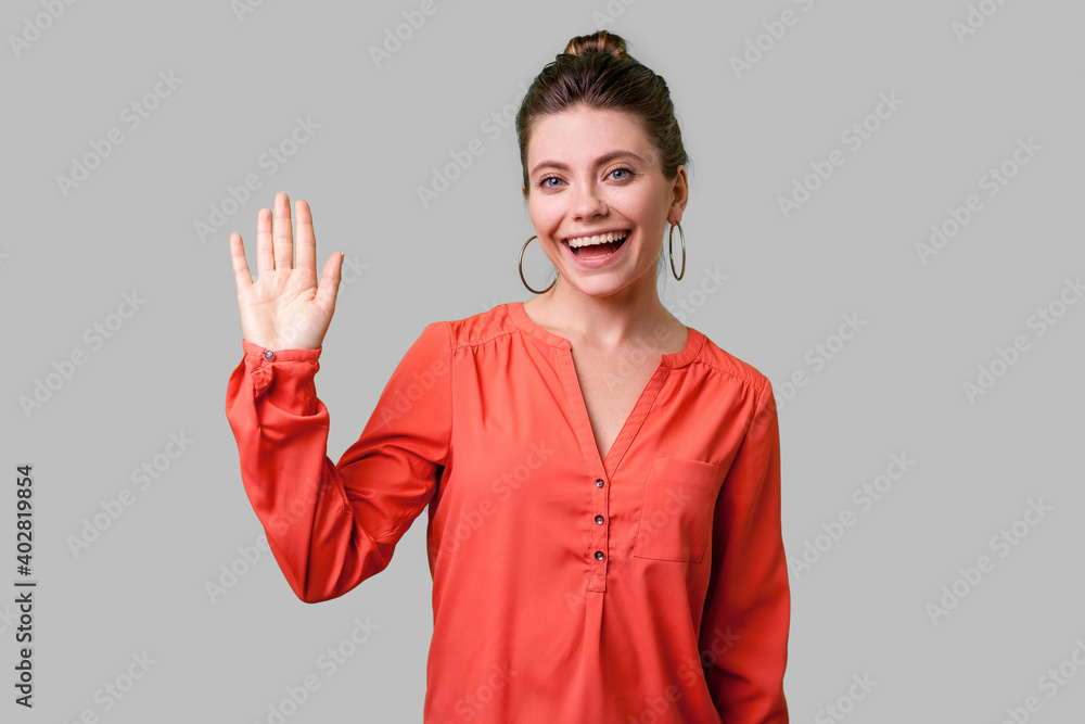 Hello! Portrait of charming woman with bun hairstyle, big earrings and in red blouse standing, waving hand and saying Hi to camera with toothy smile. indoor studio shot isolated on gray background Hello! Portrait of charming woman with bun hairstyle, big earrings and in red blouse standing, waving hand and saying Hi to camera with toothy smile. indoor studio shot isolated on gray background