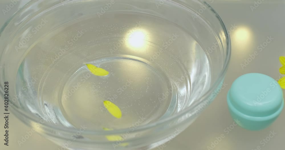 Yellow flower petals fall into a large glass bowl into the water, next to it lies a small jar of face cream and a whole flower. The camera moves slowly on the slider. Yellow flower petals fall into a large glass bowl into the water, next to it lies a small jar of face cream and a whole flower. The camera moves slowly on the slider.