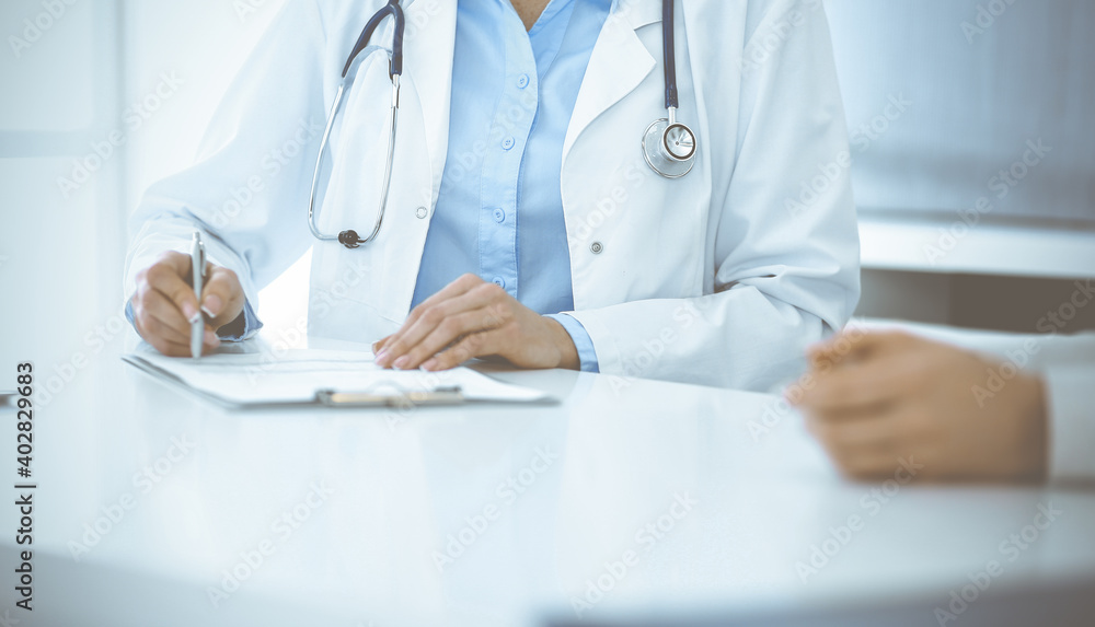 Unknown woman-doctor and female patient sitting and talking at medical examination in clinic, close-up. Therapist wearing blue blouse is filling up medication history record. Medicine concept Unknown woman-doctor and female patient sitting and talking at medical examination in clinic, close-up. Therapist wearing blue blouse is filling up medication history record. Medicine concept