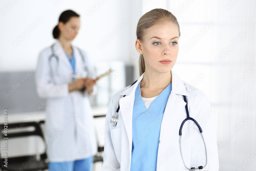 Woman - doctor standing in clinic with colleague at the background. Physician at work. Medicine and health care concept Woman - doctor standing in clinic with colleague at the background. Physician at work. Medicine and health care concept