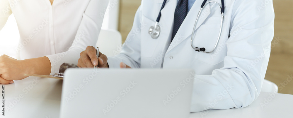 Unknown doctor and patient discussing something while sitting at the desk in sunny clinic, close-up Unknown doctor and patient discussing something while sitting at the desk in sunny clinic, close-up