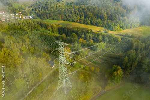 Aerial view of the high voltage power lines and high voltage electric transmission on the terrain in clouds surrounded by trees at sunlight