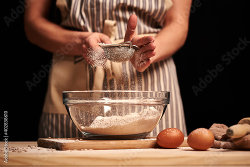 Female baker hands making dough for bread with an apron. Natural homemade ingredients. Dark background, brown color grading.
