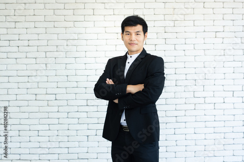 portrait of young asian businessman in suit arm crossed standing and smile with white brick background.