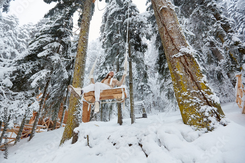 Young girl swinging on a long swing on snow forest mountain view. Swinging between tall pines. Bottom-up view