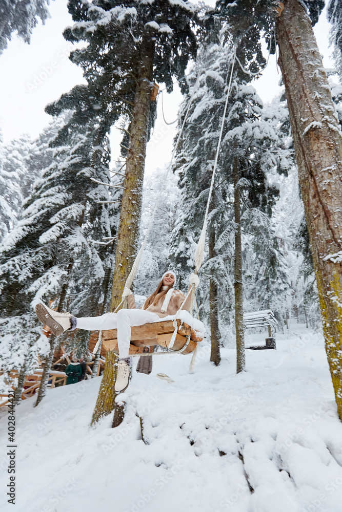 Young girl swinging on a long swing on snow forest mountain view. Swinging between tall pines. Bottom-up view Young girl swinging on a long swing on snow forest mountain view. Swinging between tall pines. Bottom-up view