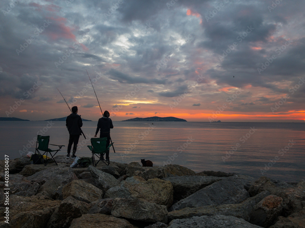 Two fisherman with chairs and rods standing on rocks during sunset and trying to catch some fishes. A cat lies beside them and waiting for food. Cloudy and dramatic sky background with some islands